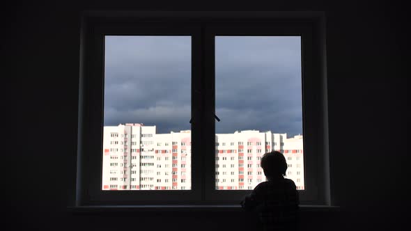 A Lonely Child Looks Out the Window of His House in an Apartment. alt