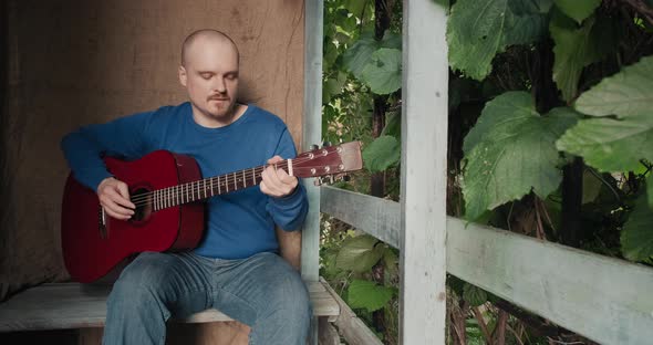 Man with an Acoustic Guitar is Sitting on Porch of House Playing an Instrument alt