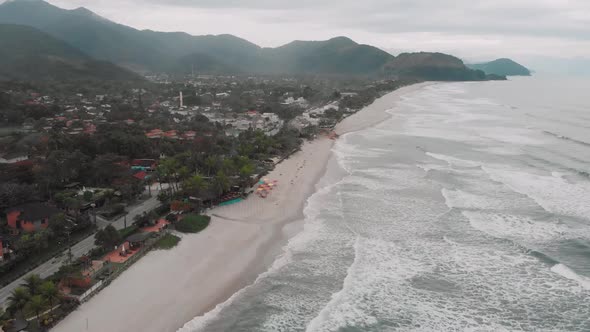 Lateral drone footage of the beach, mountain and houses in front of the sea, cloudy day, waves, land alt