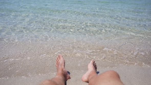 Closeup of Men's Feet Lying on the Seashore Small Waves Hitting Their Feet on the Shore in Slow alt