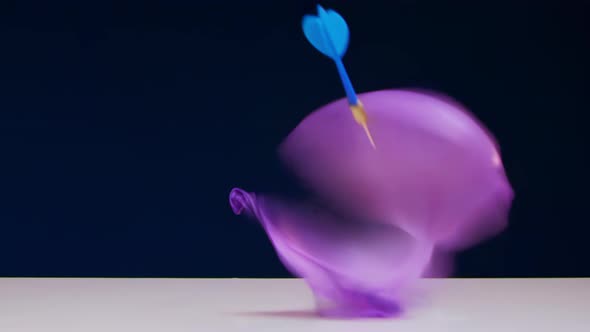 Red Dart Blows Up Green Balloon Lying on White Table Against Blue Background