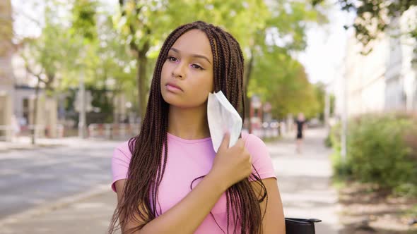 A Young Black Woman Takes Off a Face Mask and Looks at the Camera in the Street in an Urban Area alt