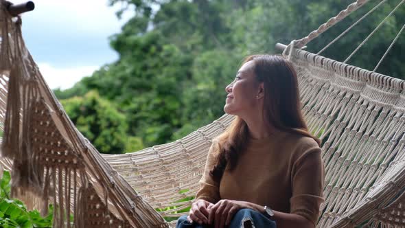 A young asian woman sitting and relaxing on hammock while looking at a beautiful nature view