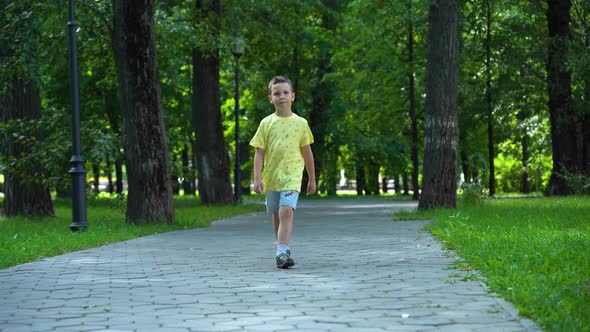 The boy is walking in the park among the green trees in the background alt