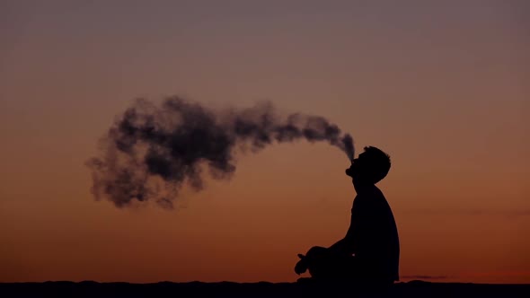 Guy Sits on the Sand and Smokes an Electronic Cigarette Against the Sunset. Silhouette alt
