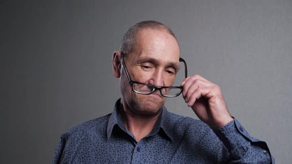 Senior Man Takes Off Glasses Rubbing Eyes on Grey Background alt