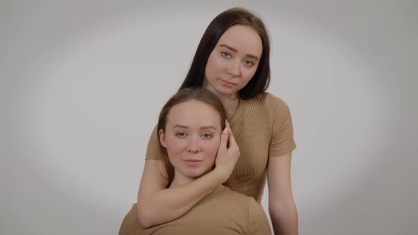 Front View Young Woman Touching Face of Twin Sister Posing at Grey Background alt