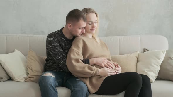 Young happy family is waiting for birth of baby sitting on sofa and hugging