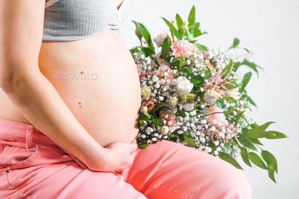 Pregnant woman with bouquet of flowers. Girl in gray top, pink pants holds hands on naked belly.  - Stock Photo - Images