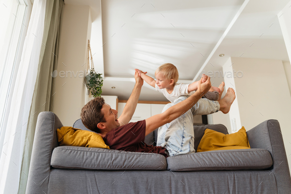 Side view of dad enjoying time with his baby son lifting him up on his legs laying on sofa