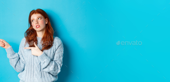 Annoyed teenage redhead girl roll eyes, pointing fingers left at something boring or lame, standing