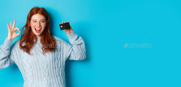 Happy redhead girl in sweater showing credit card and okay sign, recommending bank offer, standing