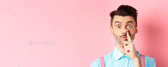 Close-up of funny caucasian man picking nose, staring silly at camera, standing on pink background