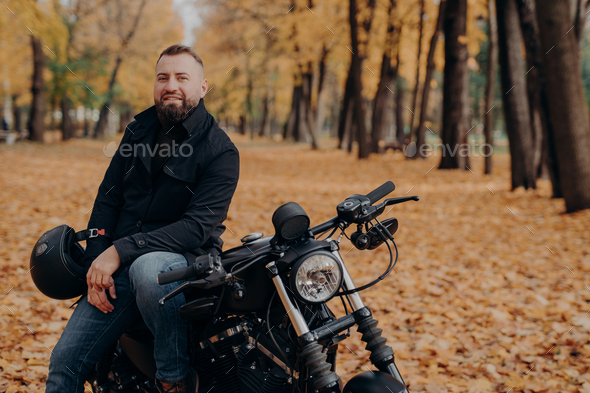 Bearded male motorcyclist rides black bike, holds helmet, has travel on his own transport