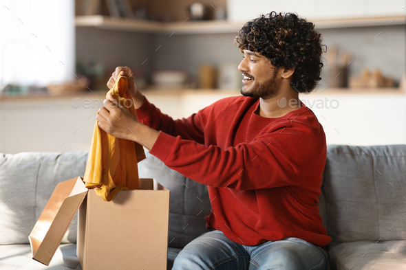 Happy Young Indian Guy Unboxing Parcel With New Clothes At Home