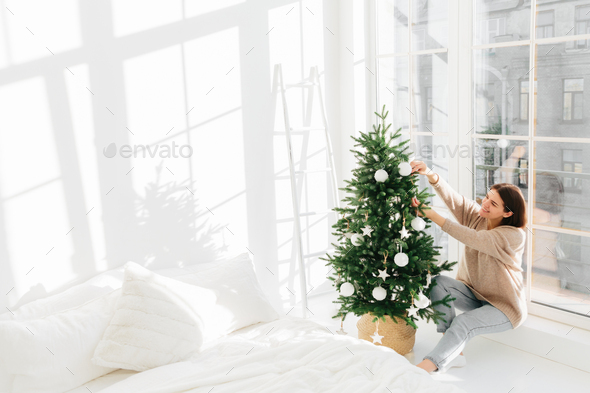 Positive young female poses near big window in cozy bedroom, enjoys decorating Christmas tree - Stock Photo - Images