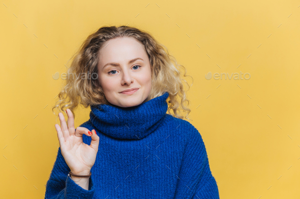 Delighted attractive young female with blonde curly hair, satisfied expression, shows ok sign - Stock Photo - Images