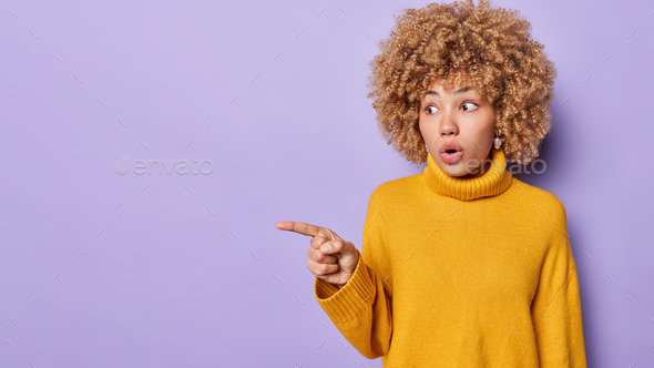 Photo of stupefiied Caucasian girl stares at camera, keeps jaw dropped, points with index finger at