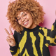 Beautiful curly haired woman smiles gladfully shows peace sign feels carefree keeps arm outstreched - PhotoDune Item for Sale