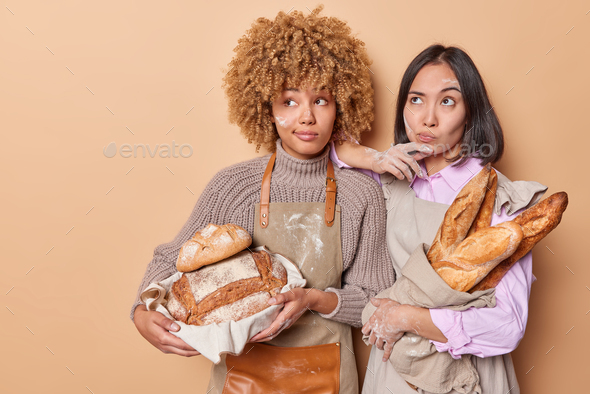 Pensive female bakers work in bakery shop holds assortment of freshly baked bread focused aside