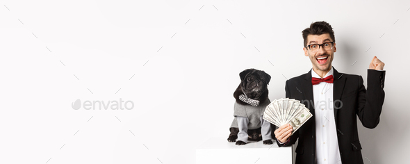 Happy young man in suit earn money with his dog. Guy rejoicing, holding dollars, black pug in
