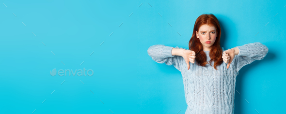 Disappointed redhead girl in sweater showing thumbs-down, judging bad product, disagree and dislike