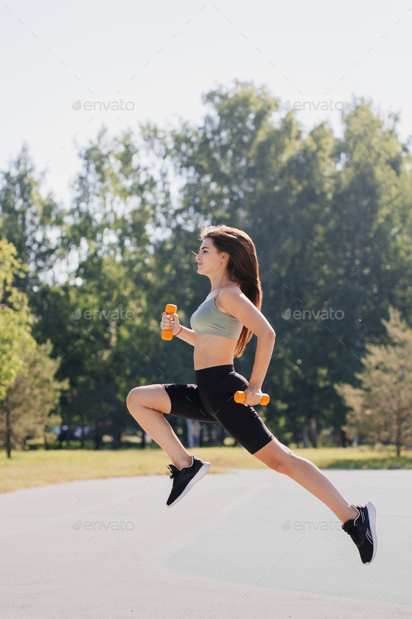Vertical shot of brunette hispanic young woman jumps high holds dumbbells against blurry park.