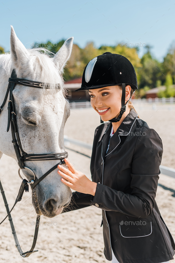 attractive smiling female equestrian standing near horse at horse club
