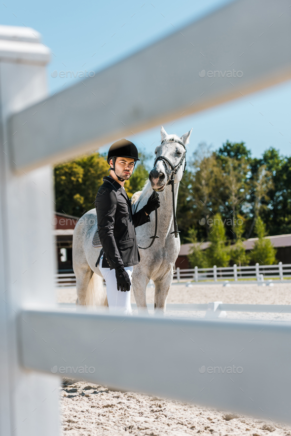 view through fence of handsome male equestrian standing near horse at horse club