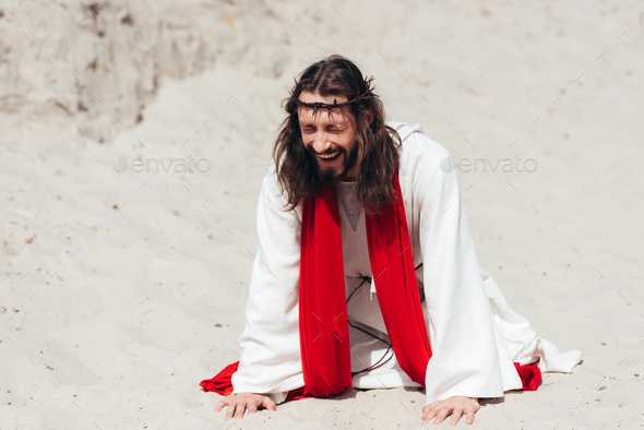laughing Jesus in robe, red sash and crown of thorns standing on knees and touching sand with hands - Stock Photo - Images