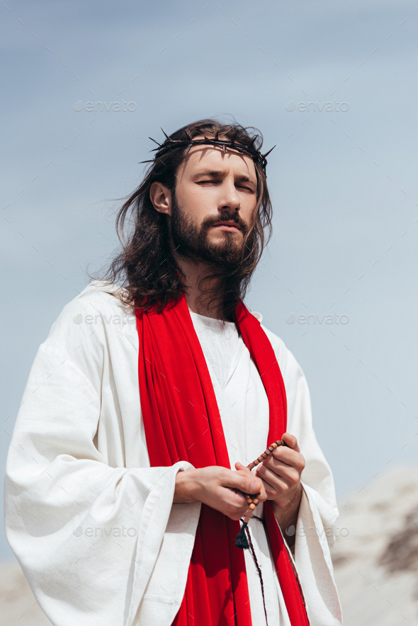 Jesus in robe, red sash and crown of thorns holding rosary and praying with  closed eyes in desert Stock Photo by LightFieldStudios