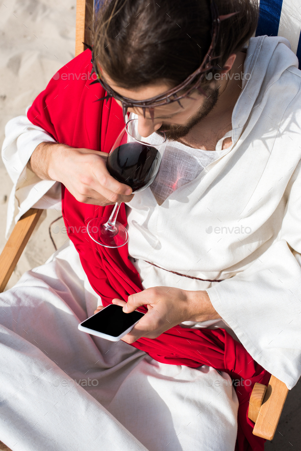 high angle view of Jesus resting on sun lounger, drinking wine and using smartphone with blank