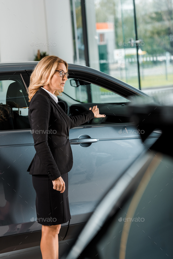 attractive female car dealer pointing at car in showroom