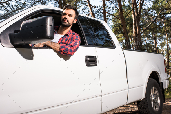 serious young handsome man sitting in pick up car and looking at wing mirror in forest