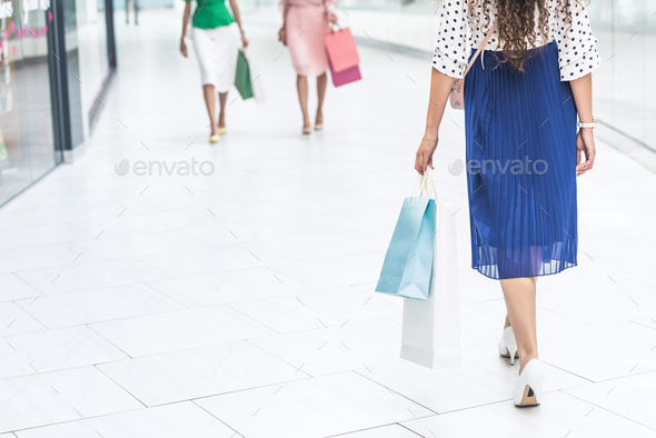 low section of stylish girls in skirts and high heeled shoes walking with shopping bags in mall