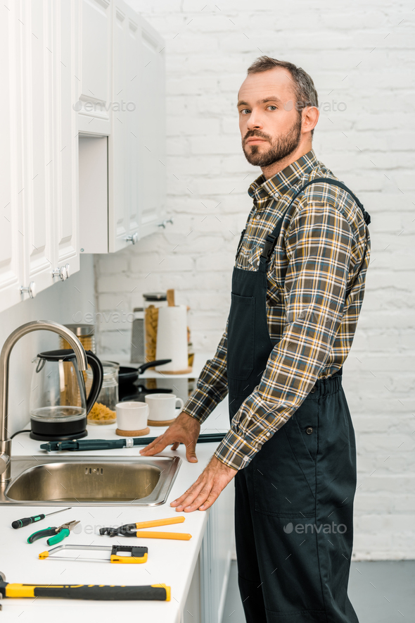 handsome plumber standing near broken sink in kitchen and looking at camera