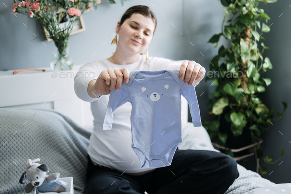 Pregnant woman with big belly holding bodysuit and sorting clothes for future. How to Organize - Stock Photo - Images
