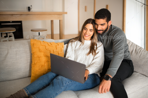 Smiling international millennial couple sit on sofa, hugging, looking at  laptop, have video call in Stock Photo by Prostock-studio