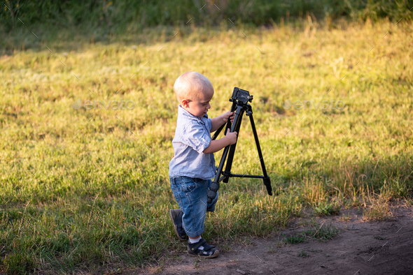 Little boy messing around, playing with tripod for camera on green meadow in the park. Hobbies