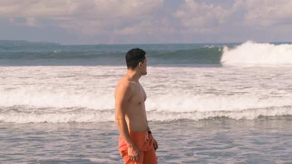 Young Athletic Tanned Millennial Man with a Bare Torso in Orange Shorts Stands in the Indian Ocean