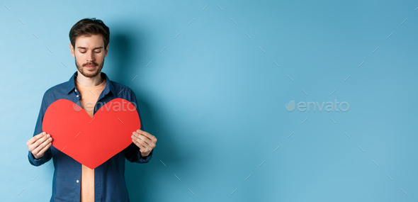 Lonely guy looking sad at valentines red heart with sad face, standing over blue background