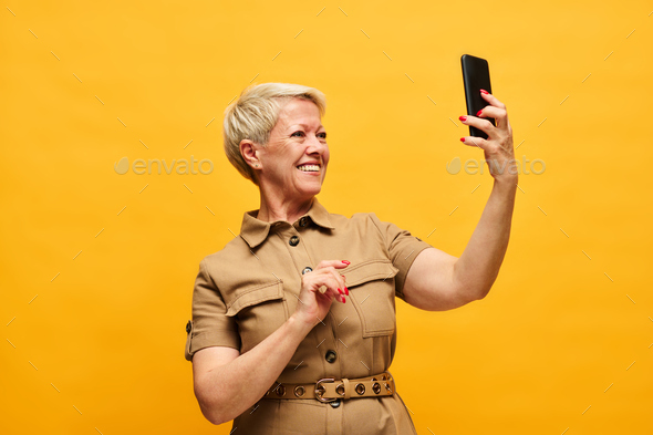 Cheerful blond woman in stylish beige dress looking at smartphone screen - Stock Photo - Images