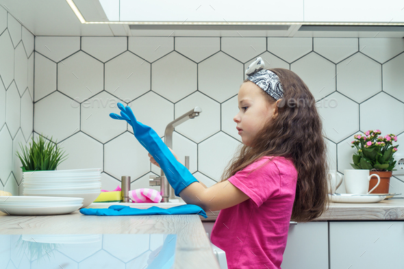 Nice little girl putting on blue household gloves for washing up dish. Home cleaning, personal