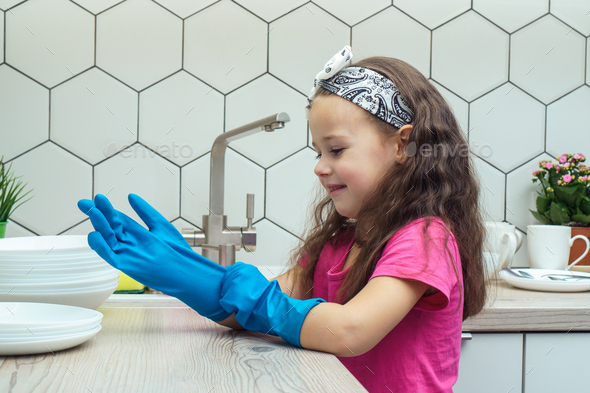 Happy little girl putting on blue household gloves for washing up dish in kitchen sink. Home