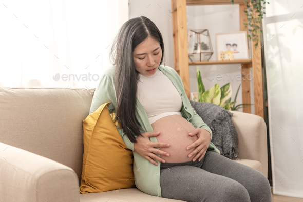 Asian young pregnant woman suffering belly ache sitting on a couch in the living room at home.