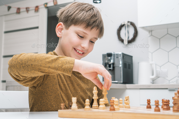 Smiling little boy joying playing chess on wooden board on table in kitchen. Young grand master