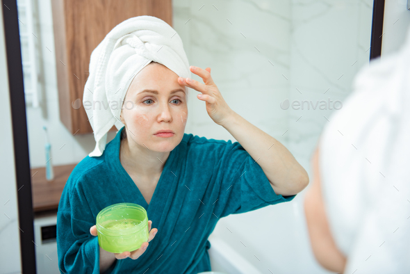 Young woman applying anti-wrinkle cream standing behind mirror in home bathroom