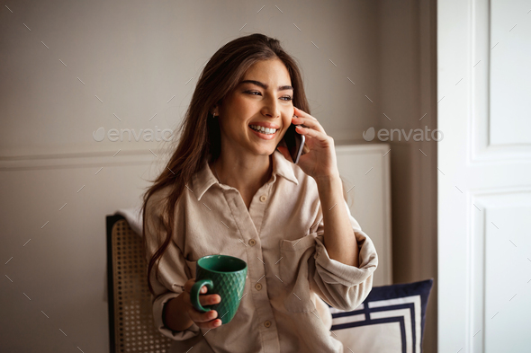 Cheerful millennial mixed race female calls by phone, talks, sits on chair, looks at empty space - Stock Photo - Images