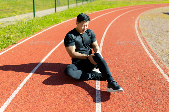 Sports injury. A young Asian sportsman, athlete, runner sits on a stadium treadmill and holds his - Stock Photo - Images