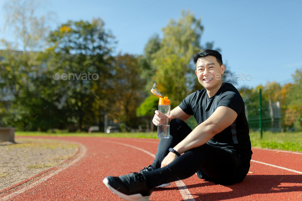 Young asian sportsman sitting in stadium on treadmill and resting after jogging, exercise. He holds - Stock Photo - Images